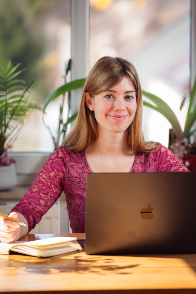 Rebecca Copywriting sitzt vor ihrem Mac am Tisch und lächelt in die Kamera